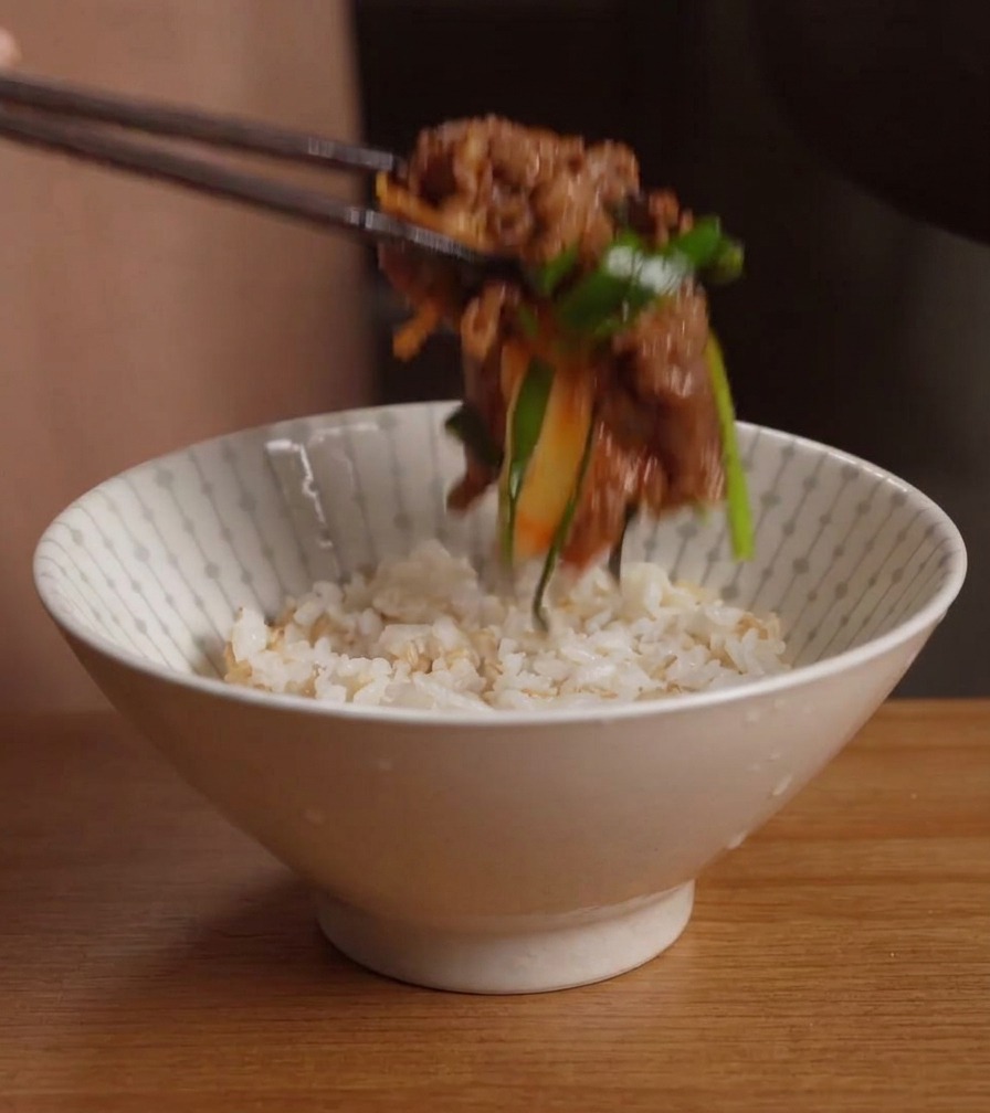 Essstäbchen setzen eine Portion der Rindfleisch-Kimchi-Pfanne auf eine weiße Schale, die mit fertigem Reis gefüllt ist.