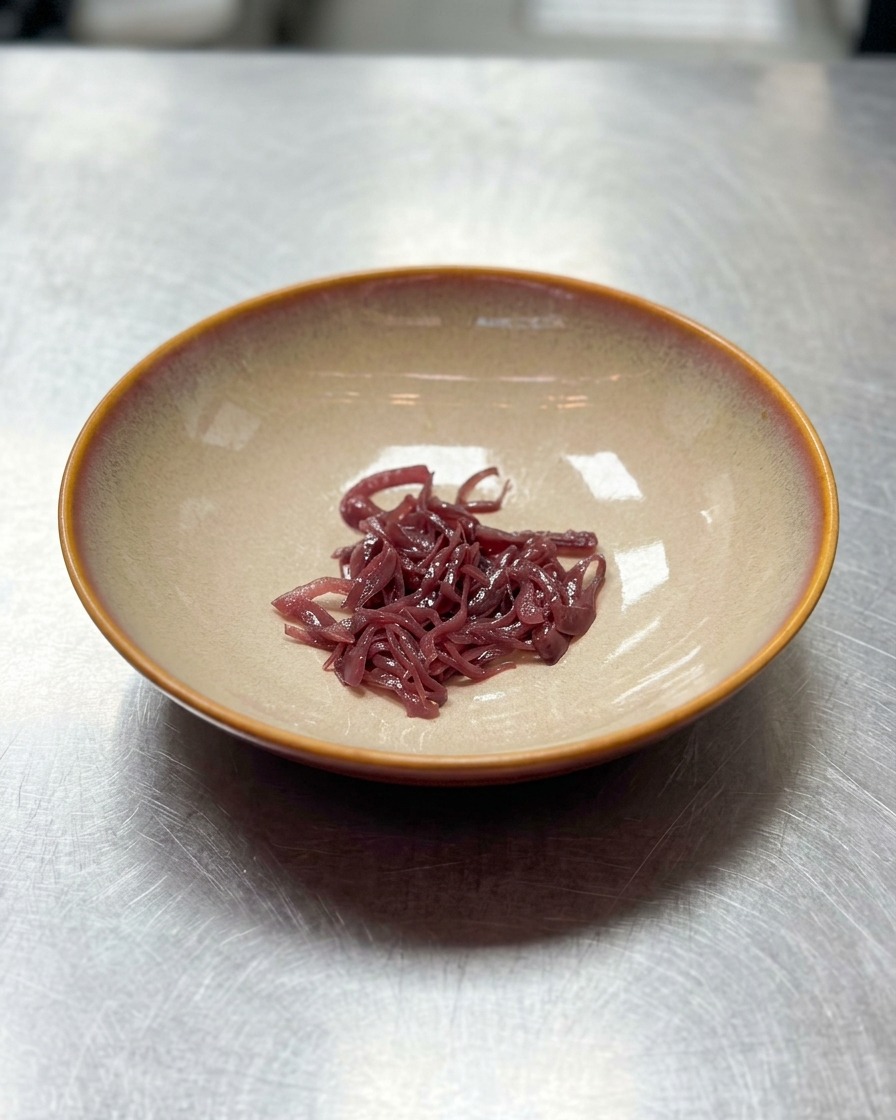 Ein kleiner Haufen in feine Streifen geschnittener roter Zwiebeln in der Mitte einer leeren, breiten, beigefarbenen Keramikschüssel.