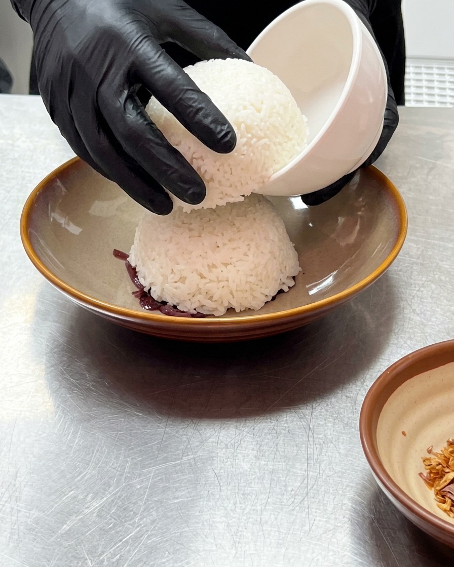 Hände in Handschuhen heben eine kleine weiße Schüssel an, um eine Kuppel aus gedämpftem weißem Reis auf den roten Zwiebeln freizulegen.