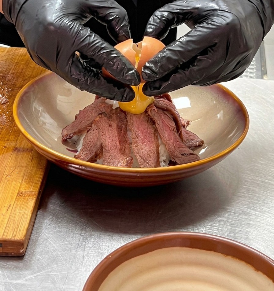 Zwei Hände in Handschuhen schlagen vorsichtig ein weiches Onsen-Ei über der Spitze des Rindfleisch-Berges auf.