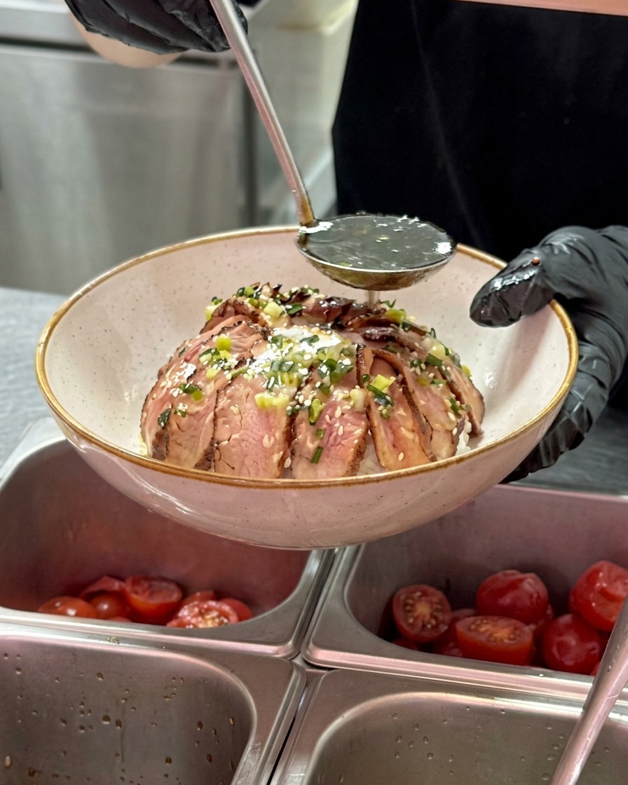 Eine Metallkelle gießt eine dunkle, glänzende Sauce über eine Schüssel mit Rindfleisch, das um ein Ei herum arrangiert ist, wobei kleine Frühlingszwiebelstücke im Öl sichtbar sind.