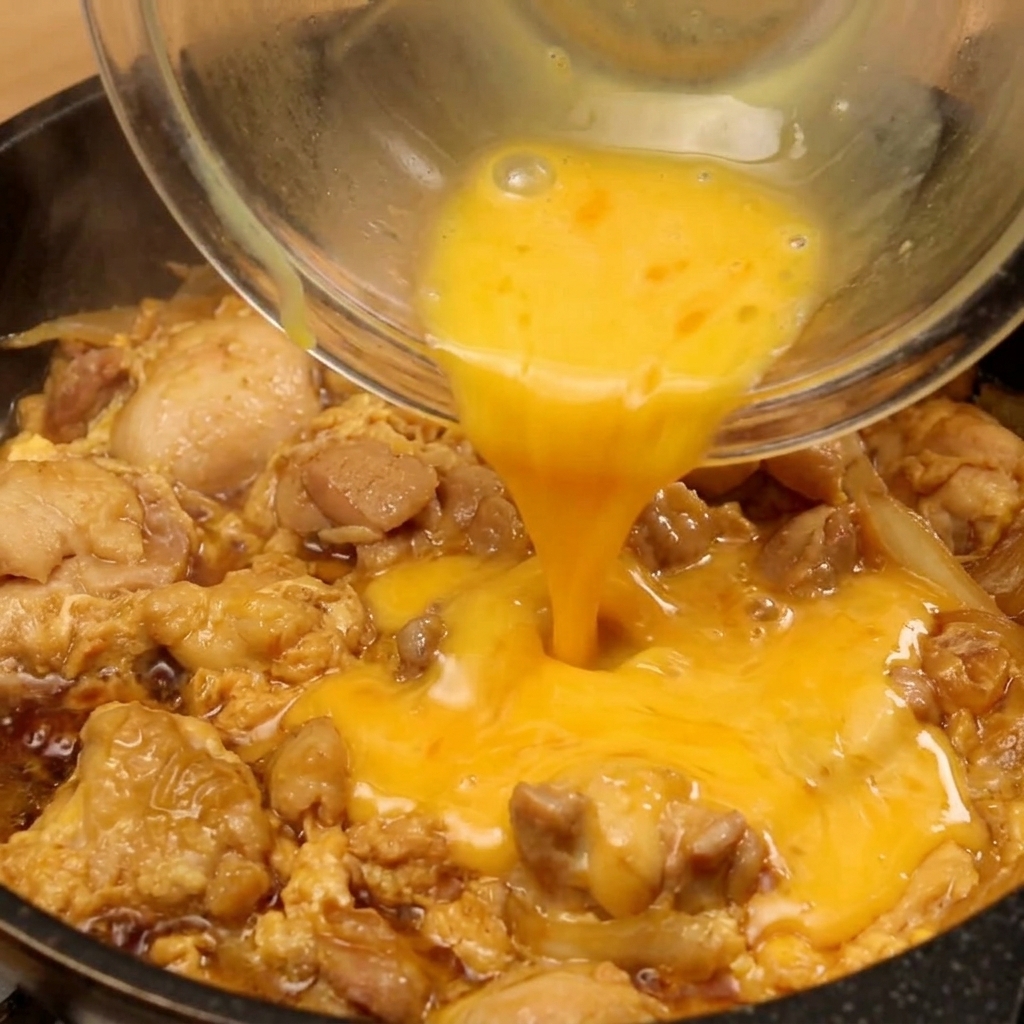Das restliche verquirlte Ei wird aus einer Glasschale in die Mitte der Pfanne gegossen, in der Hähnchen und Zwiebeln in dunkler Sauce köcheln.