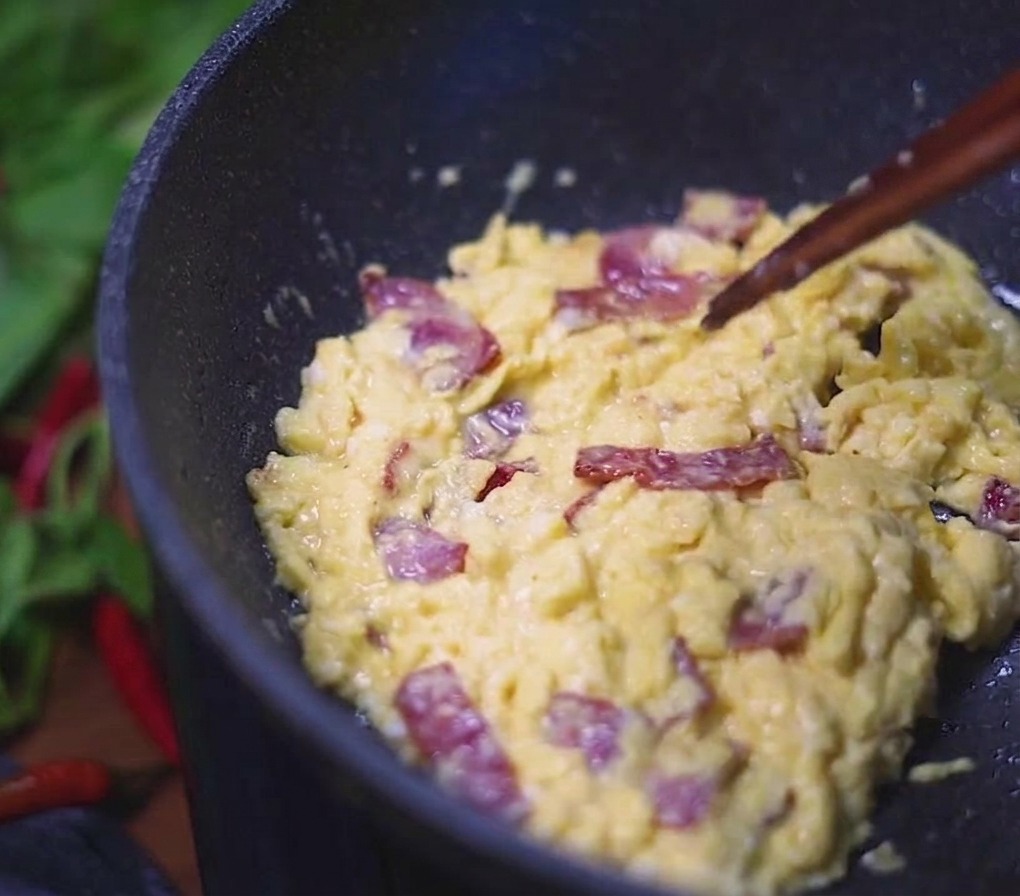 Cremiges Rührei vermischt mit herzhaften Speckstücken wird mit Stäbchen in einer dunklen Pfanne verrührt.
