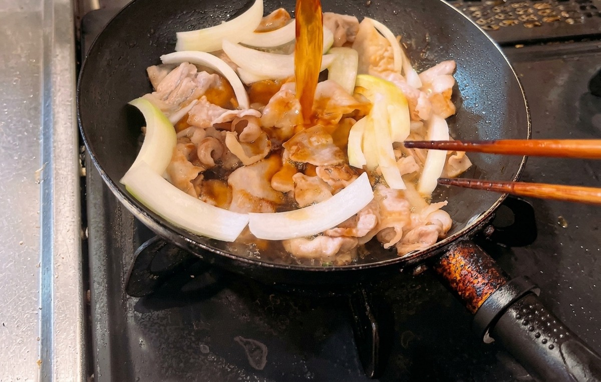 Eine bernsteinfarbene Sauce wird in eine Bratpfanne gegossen, die gebratenen Schweinebauch und rohe weiße Zwiebelstreifen enthält.