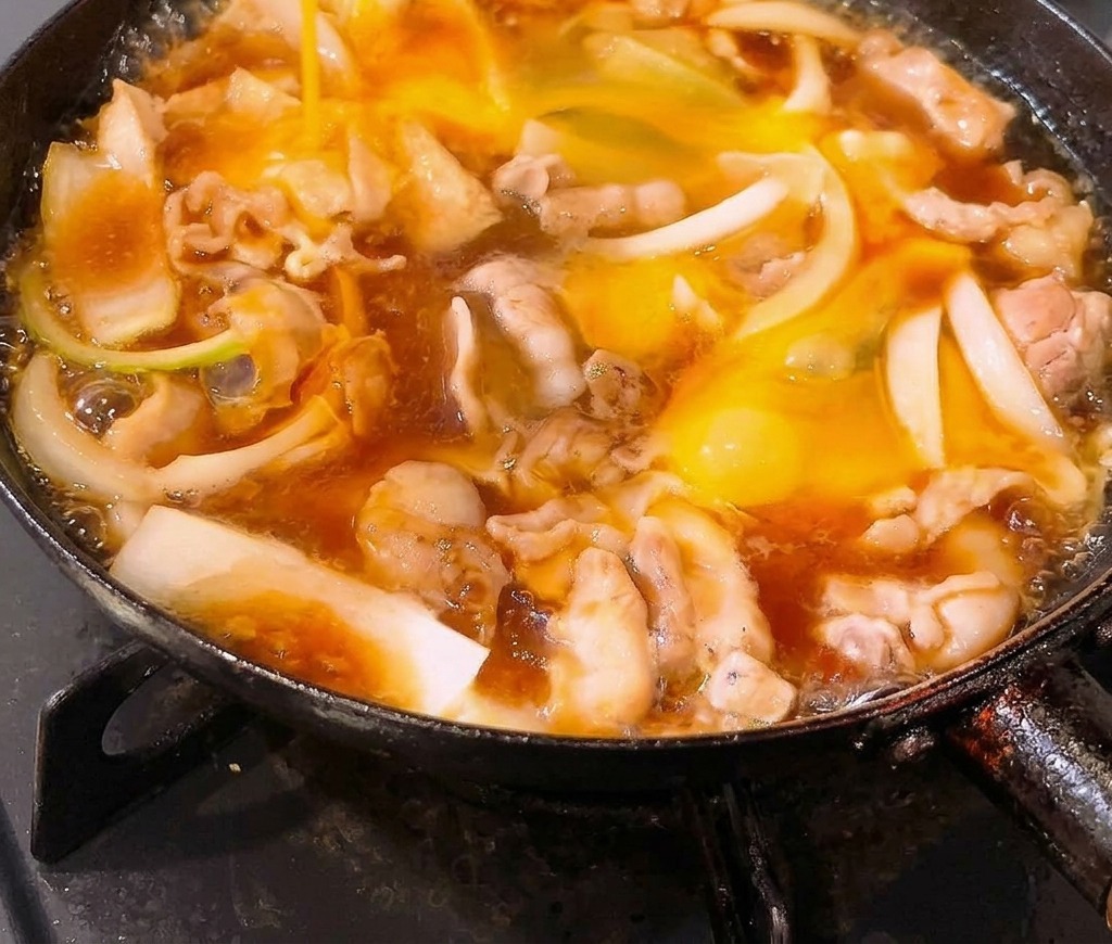 Verquirlte Eier werden in eine Bratpfanne gegossen, die mit köchelndem Schweinebauch und Zwiebeln in einer Sauce auf Dashibasis gefüllt ist.