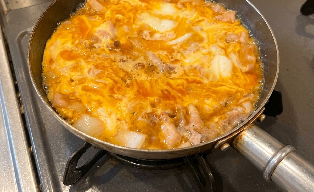 Eine Pfanne mit Tanindon köchelt auf dem Herd, wobei die gelben Eier trüb und perfekt um das Schweinefleisch und die Zwiebeln herum gestockt sind.