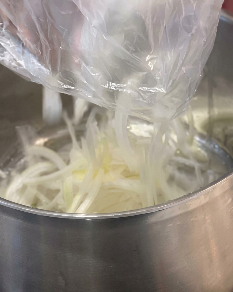 Fein geschnittene weiße Zwiebeln werden aus einer Plastiktüte in einen Edelstahltopf zu einem Stück gelber Butter gegeben.