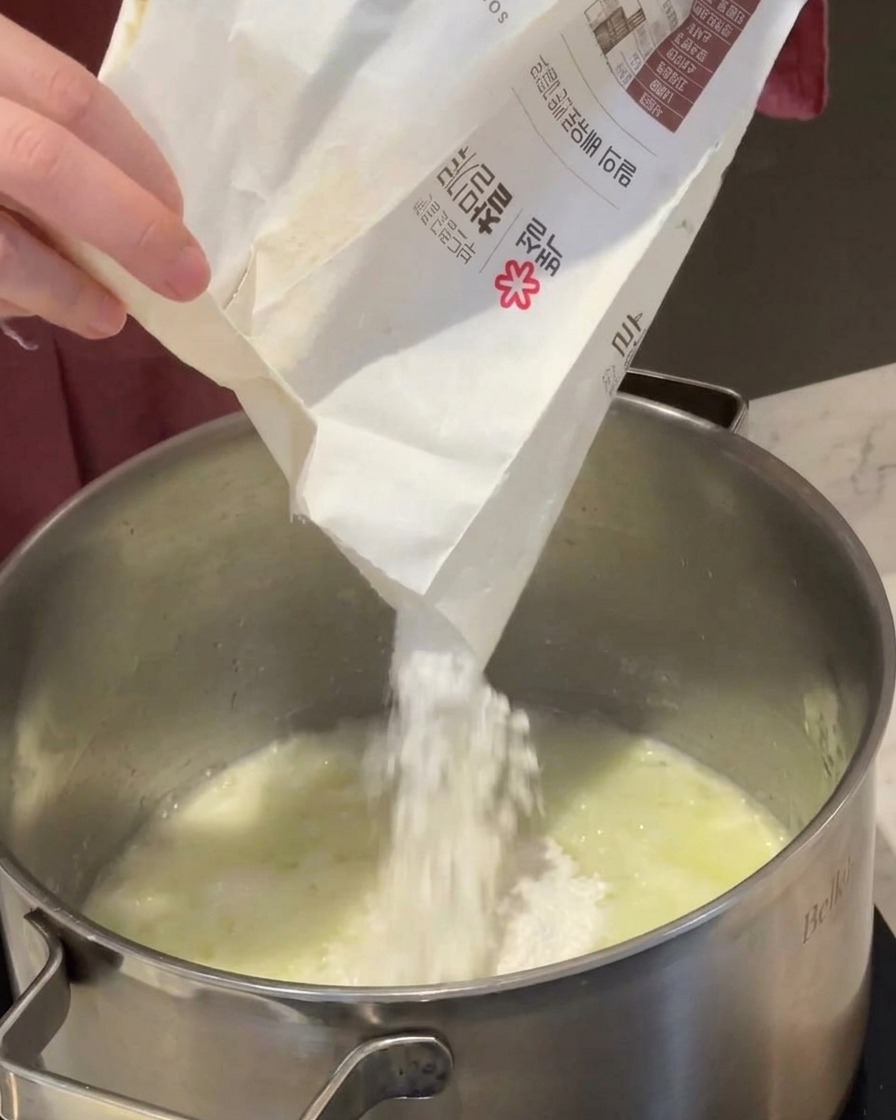 Weißes Mehl wird aus einer Papiertüte in einen Topf mit geschmolzener Butter und Zwiebeln geschüttet.