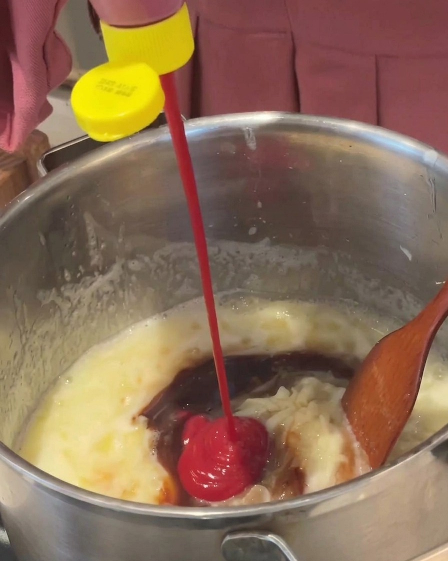 Roter Ketchup wird aus einer Flasche in eine köchelnde braune Sauce in einem Metalltopf gegeben.