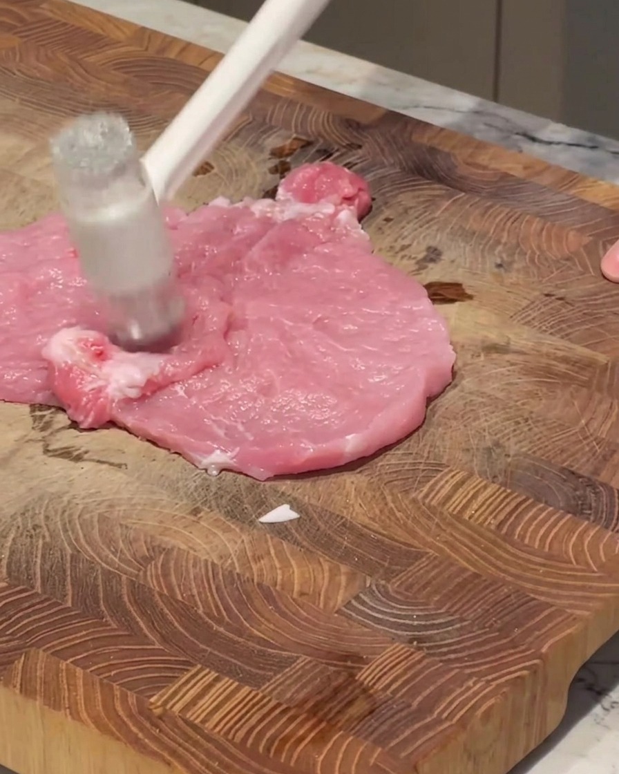 Ein weißer Fleischklopfer schlägt auf ein Stück rohes, rosa Schweinefleisch auf einem Holzbrett.