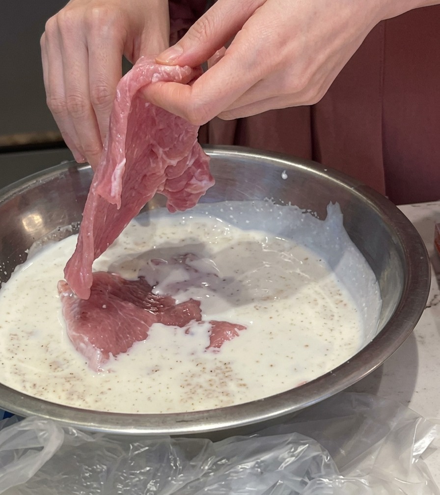 Hände legen ein dünnes Stück rohes Schweinefleisch in eine Metallschüssel mit einer weißen Milch-Knoblauch-Marinade.