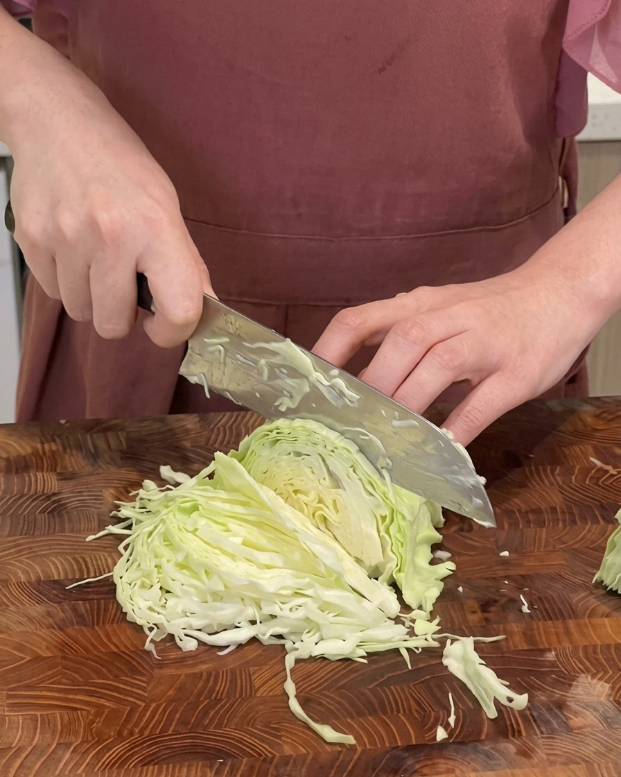 Nahaufnahme eines Kochs, der Weißkohl mit einem großen Messer auf einem Holzschneidebrett in feine Streifen schneidet.