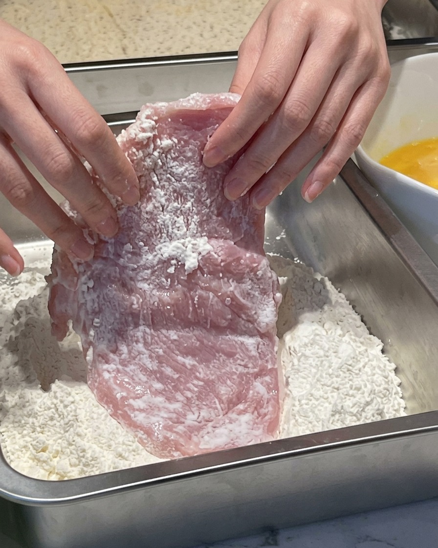 Hände drücken ein feuchtes Stück Schweinefleisch in eine Metallschale mit weißem Mehl.