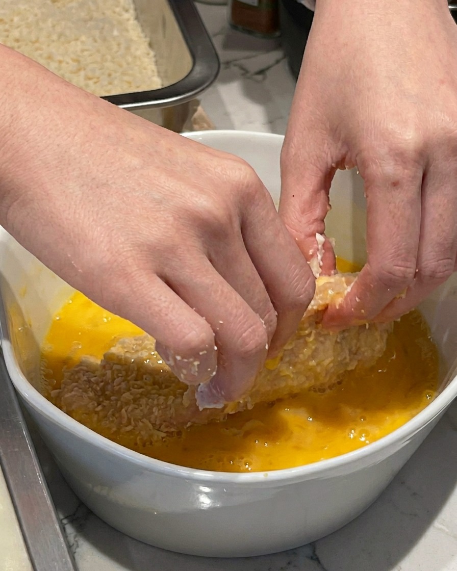 Eine Hand taucht ein mehliertes Schweineschnitzel in eine weiße Keramikschüssel mit verquirltem gelbem Ei.