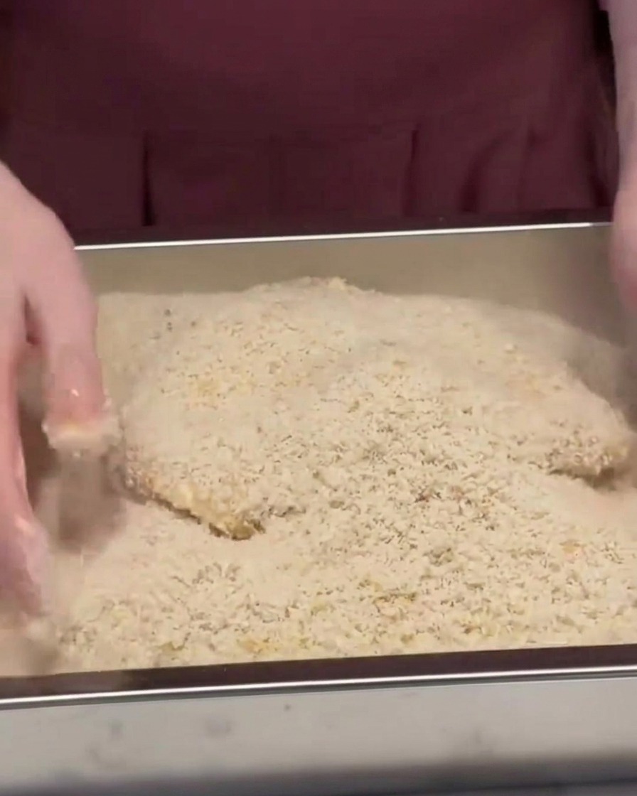 Ein Schweineschnitzel wird in einer Metallschale mit Panko-Paniermehl bedeckt und festgedrückt.