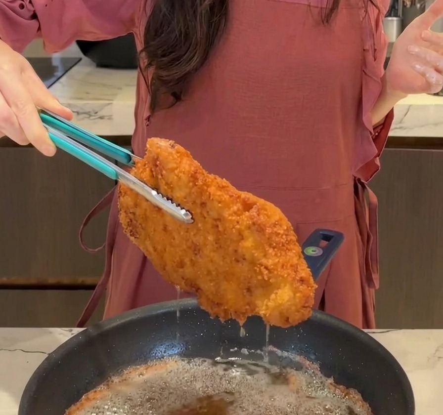 Ein großes, knuspriges, goldbraunes Tonkatsu-Schnitzel wird mit einer blauen Zange aus der Pfanne gehoben, während Öl abtropft.