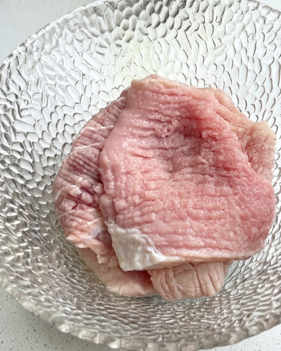 Nahaufnahme einer Glasschüssel mit rohen Schweinefleischscheiben, garniert mit gehackten Frühlingszwiebeln und flüssigen Gewürzen.