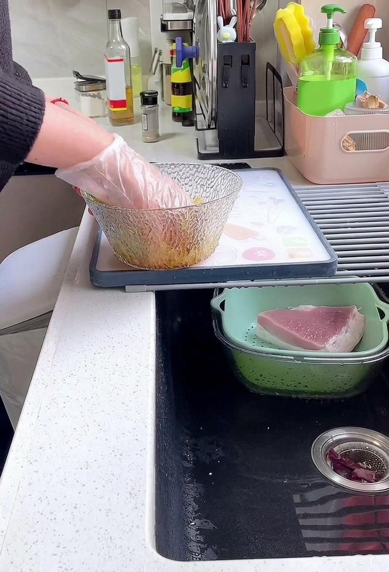 Eine Person mit einem Plastikhandschuh mischt dicke Schweinerückenscheiben mit Sojasauce, Pfeffer und Stärke in einer Glasschüssel.