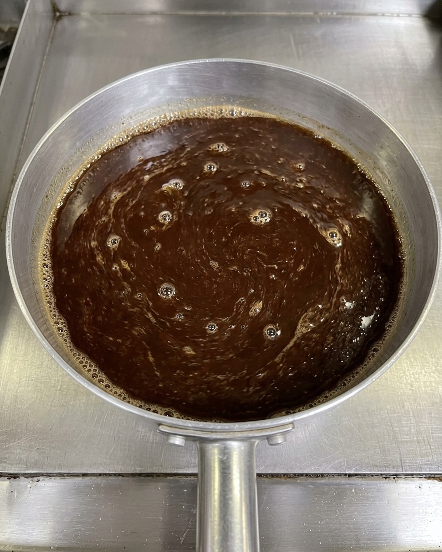 Draufsicht auf eine dunkelbraune Miso-Saucenmischung, die mit einem Paar Stäbchen in einem kleinen Aluminiumtopf umgerührt wird.