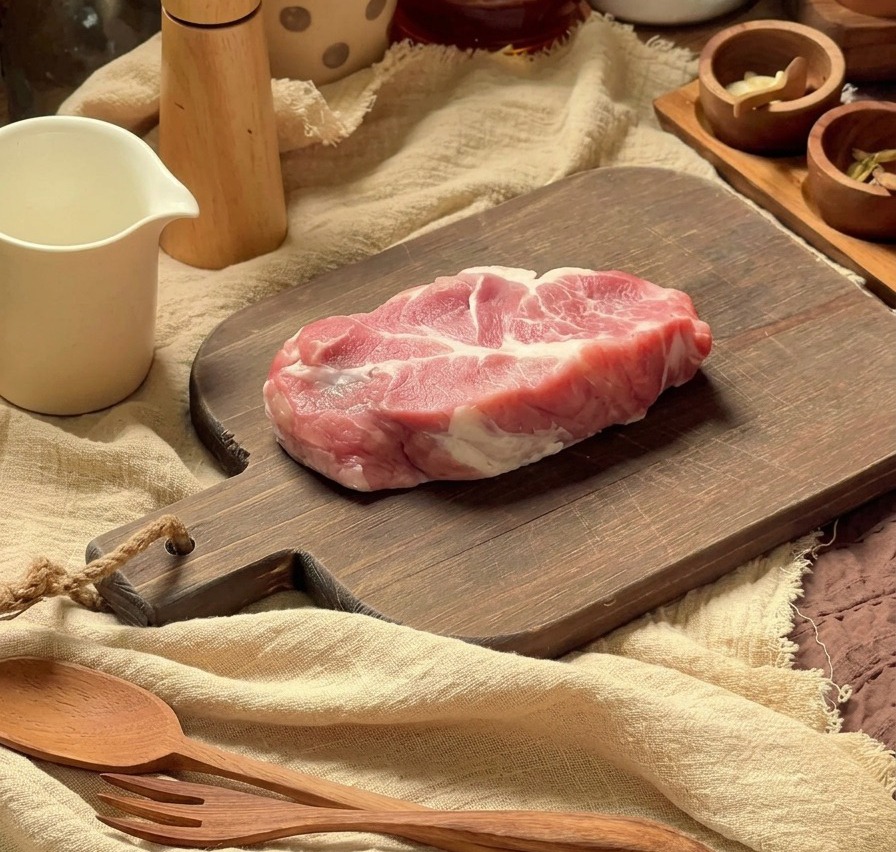 Rohes, gut marmoriertes Schweinekotelett auf einem Holzschneidebrett mit Küchenutensilien im Hintergrund.