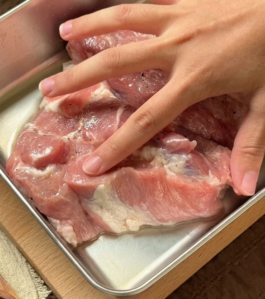 Eine Hand arbeitet eine klare, aromatische Flüssigkeit in rohe Schweinekoteletts in einer Metallschale ein.
