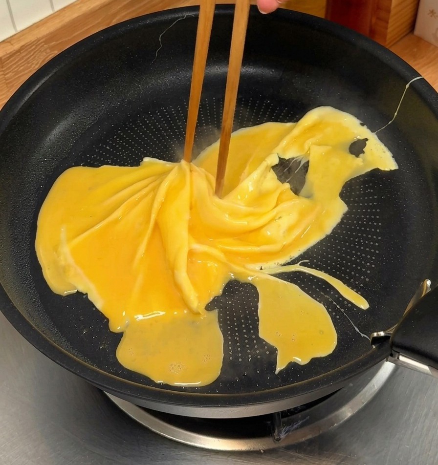 Essstäbchen drehen ein leicht gestocktes, gelbes Omelett in einer schwarzen beschichteten Pfanne, um ein Spiralmuster zu erzeugen.