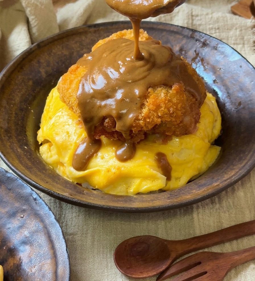 Eine dicke, glatte braune Currysauce wird mit einem Holzlöffel über ein knuspriges Schweinekotelett auf einem Tornado-Omelett gegossen.