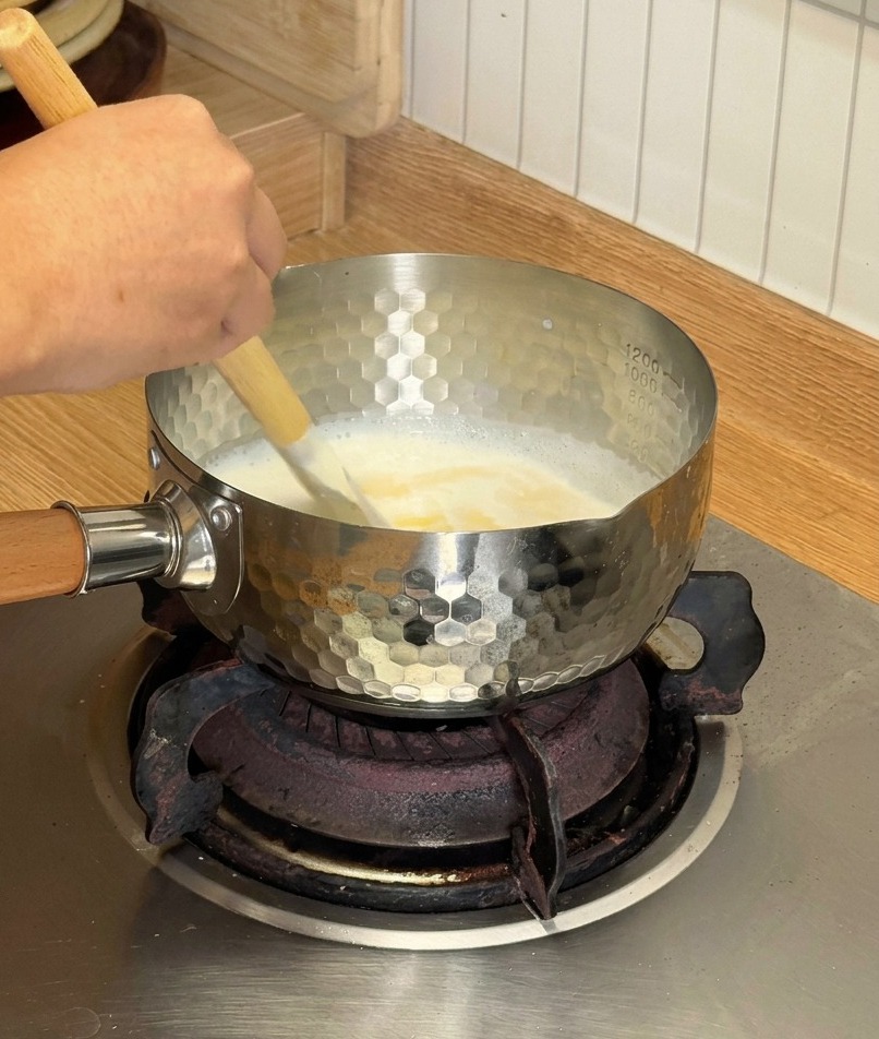 Ein Holzspatel rührt eine cremige weiße Mischung aus schmelzendem Käse und Kokosmilch in einem kleinen silbernen Topf auf dem Herd.