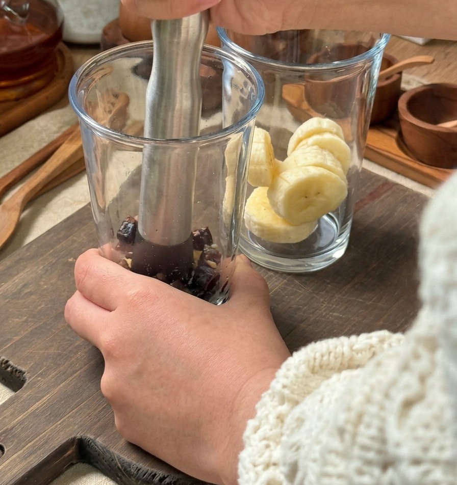 Hände zerdrücken Zutaten mit einem silbernen Stößel in einem hohen Glas, daneben steht ein weiteres Glas mit Bananenscheiben bereit.