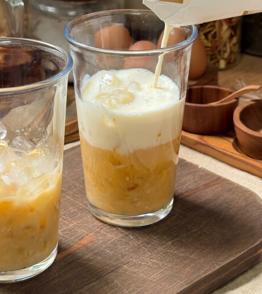 Kalte Milch wird aus einer Packung in ein hohes, mit Eiswürfeln und gelbem Bananenpüree gefülltes Glas gegossen.