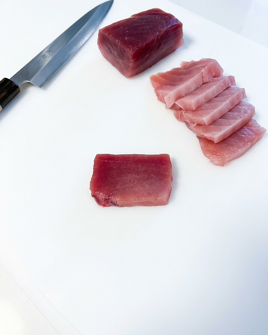 Mehrere rechteckige Scheiben von rosafarbenem, fettem Thunfisch liegen neben einem großen Thunfischblock und einem japanischen Küchenmesser auf einem weißen Brett.