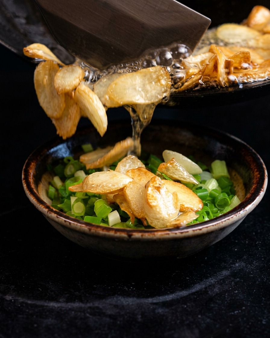 Zischendes heißes Öl und frittierte Knoblauchchips werden aus einer Pfanne in eine Schüssel mit gehackten Frühlingszwiebeln gegossen.