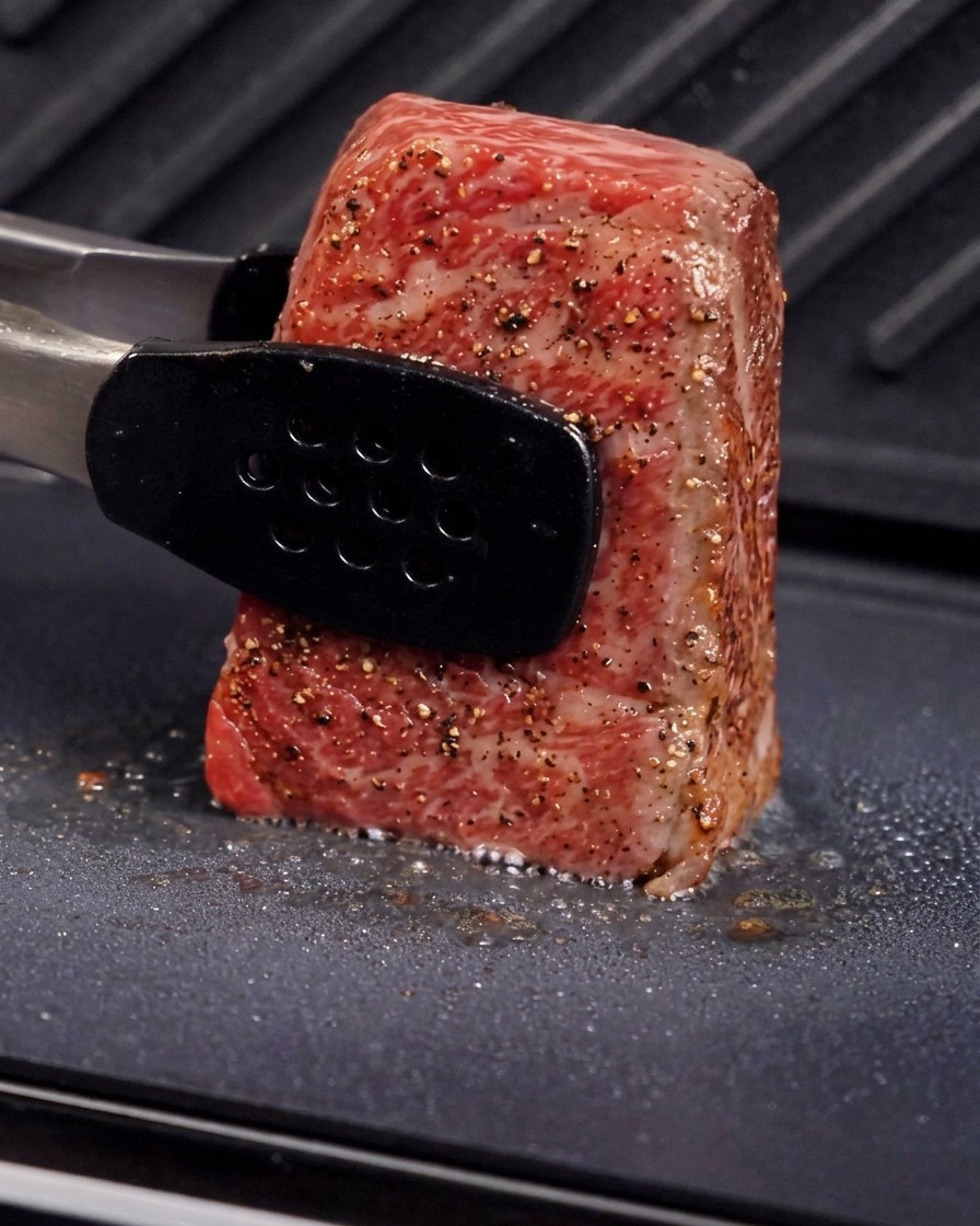 Eine Metallzange hält ein dickes Stück Fleisch aufrecht auf einer Grillplatte, um die Seite anzubraten, wobei Dampf und zischendes Öl zu sehen sind.