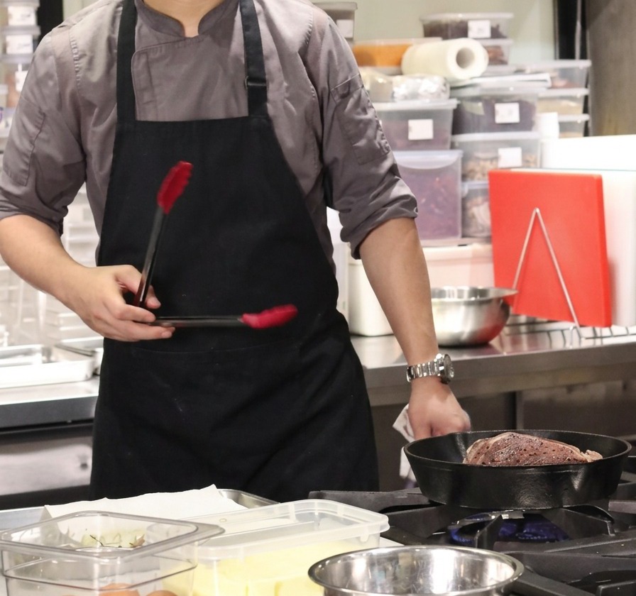 Ein Koch hält ein großes Stück Rindfleisch mit einer Zange in einer heißen gusseisernen Pfanne, um die Fettschicht anzubraten.