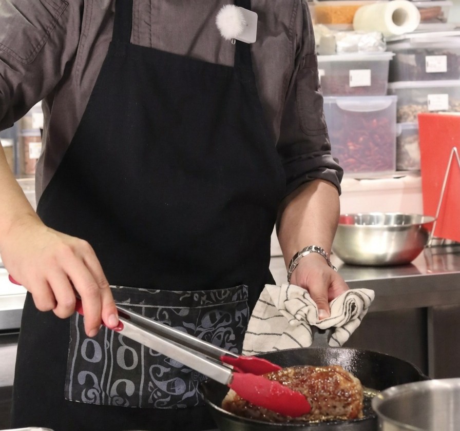 Nahaufnahme eines großen Rindfleischstücks, das vertikal mit einer rot markierten Zange in einer heißen gusseisernen Pfanne gehalten wird.