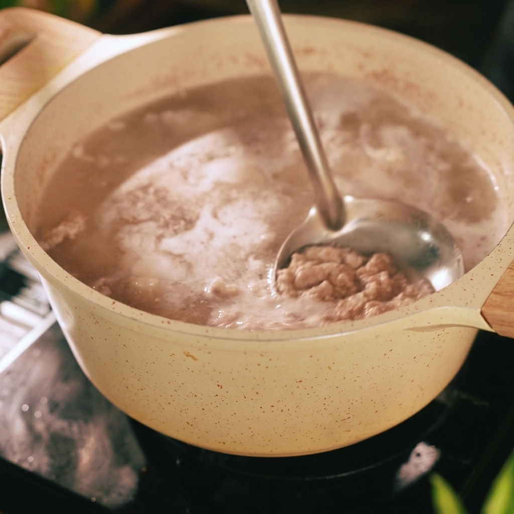 Eine Schöpfkelle schöpft grauen Schaum und Unreinheiten von der Oberfläche von kochendem Wasser in einem beigen Topf mit Schweinebauchstreifen.
