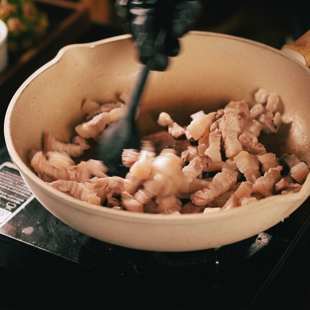Schweinebauchstreifen werden in einer beigen Pfanne mit einem schwarzen Pfannenwender angebraten, zeigen Bräunung und ausgelassenes Fett.
