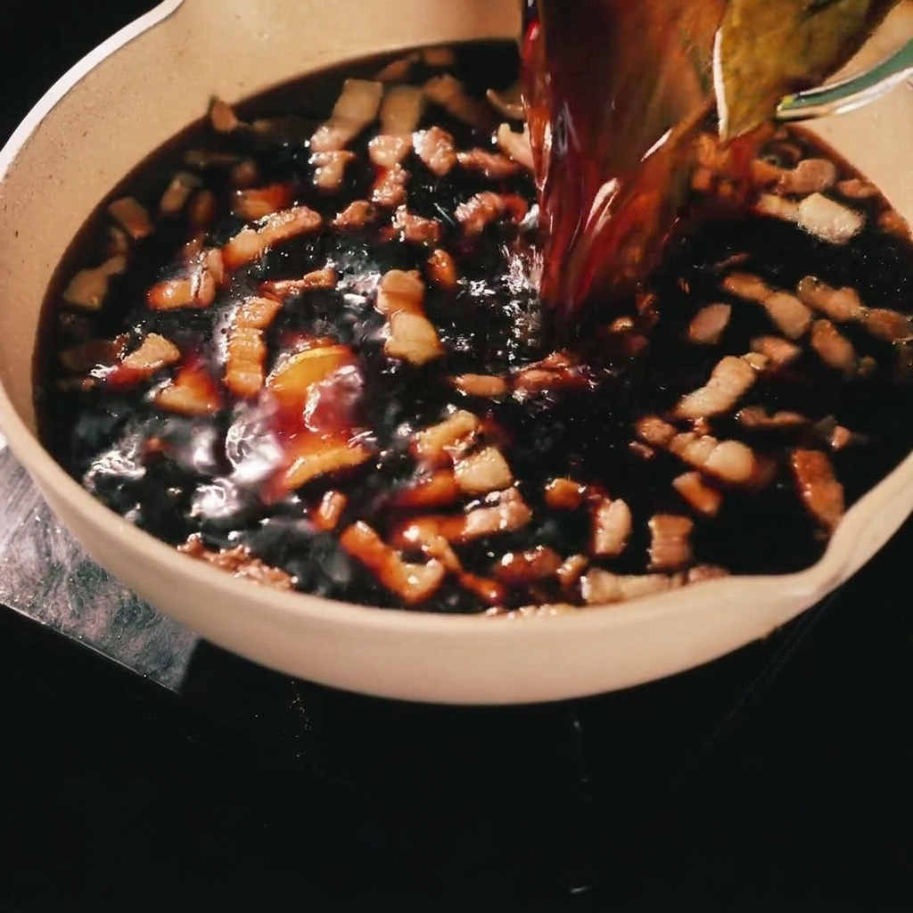 Eine dunkle, herzhafte Marinade wird aus einem Glasbehälter über angebratene Schweinebauchstreifen in einer Pfanne gegossen.