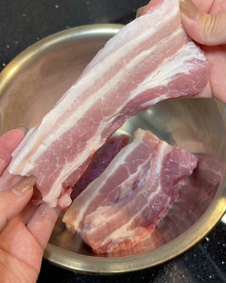 Zwei Hände halten einen langen Streifen rohen Schweinebauch ohne Schwarte über einer Edelstahlschüssel.