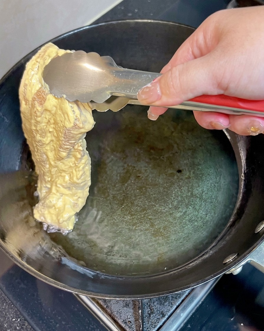 Ein Stück panierter Schweinebauch wird mit einer Metallzange vorsichtig in heißes Öl in einer Pfanne gelegt.