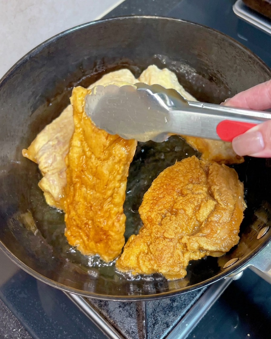 Eine Metallzange wendet ein Stück knusprigen, goldbraunen Schweinebauch in sprudelndem, heißem Öl neben anderen Stücken.