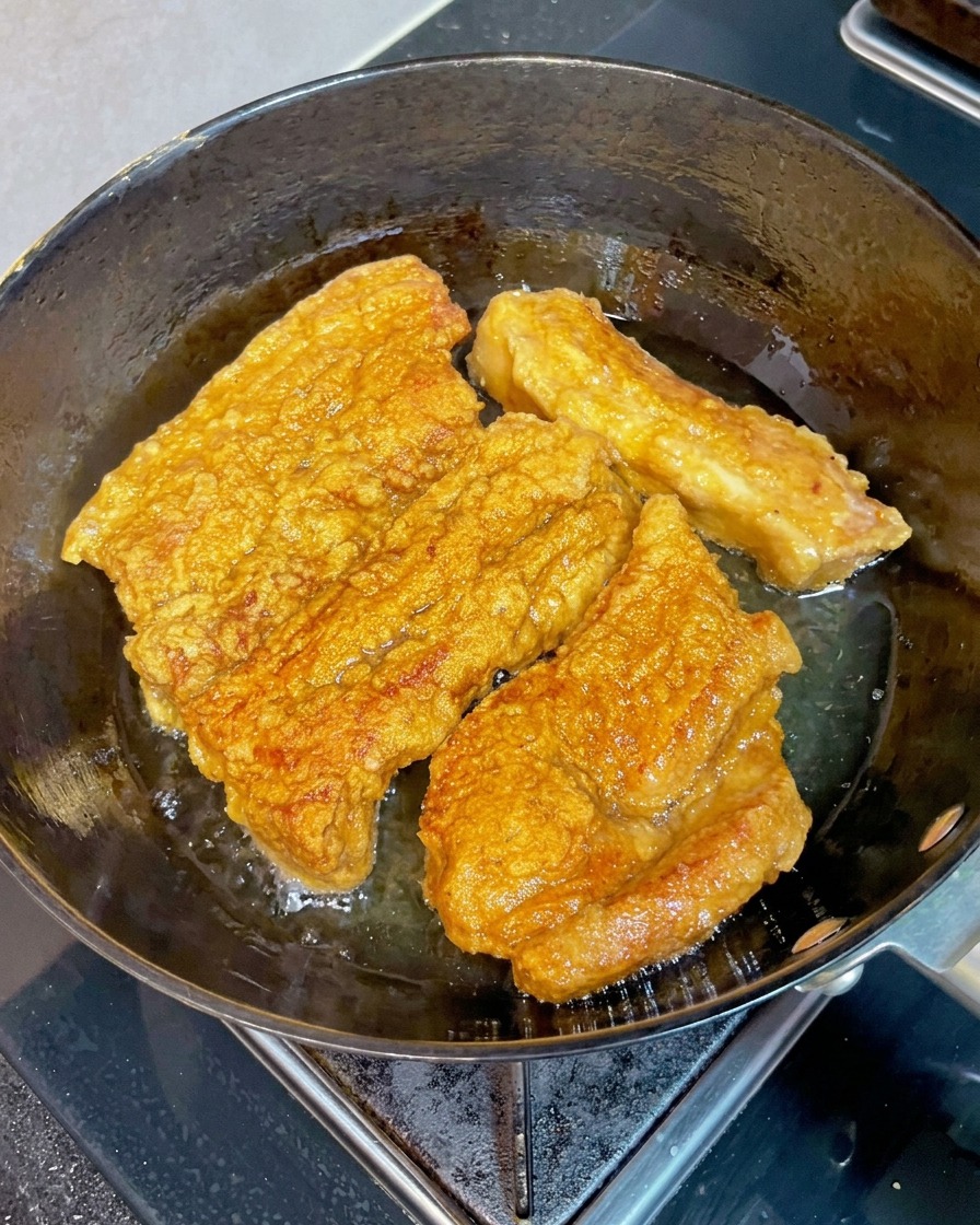 Knusprige goldbraune Schweinebauchstücke frittieren in heißem Öl, während ein Holzstäbchen in ein Stück gedrückt wird, um die Zartheit zu prüfen.