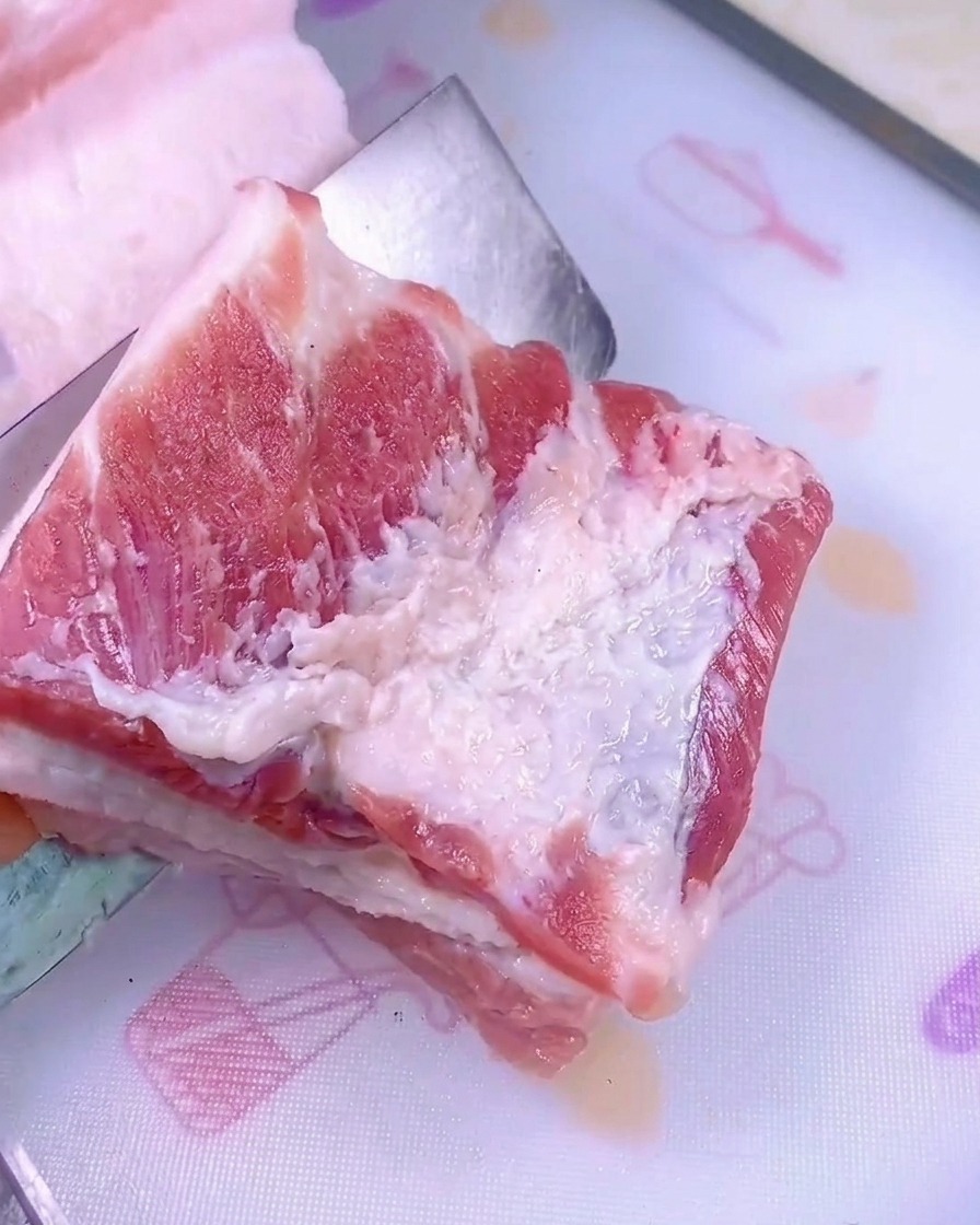 Ein scharfes Messer schneidet ein dickes Stück rohen Schweinebauch auf einem weißen Schneidebrett.
