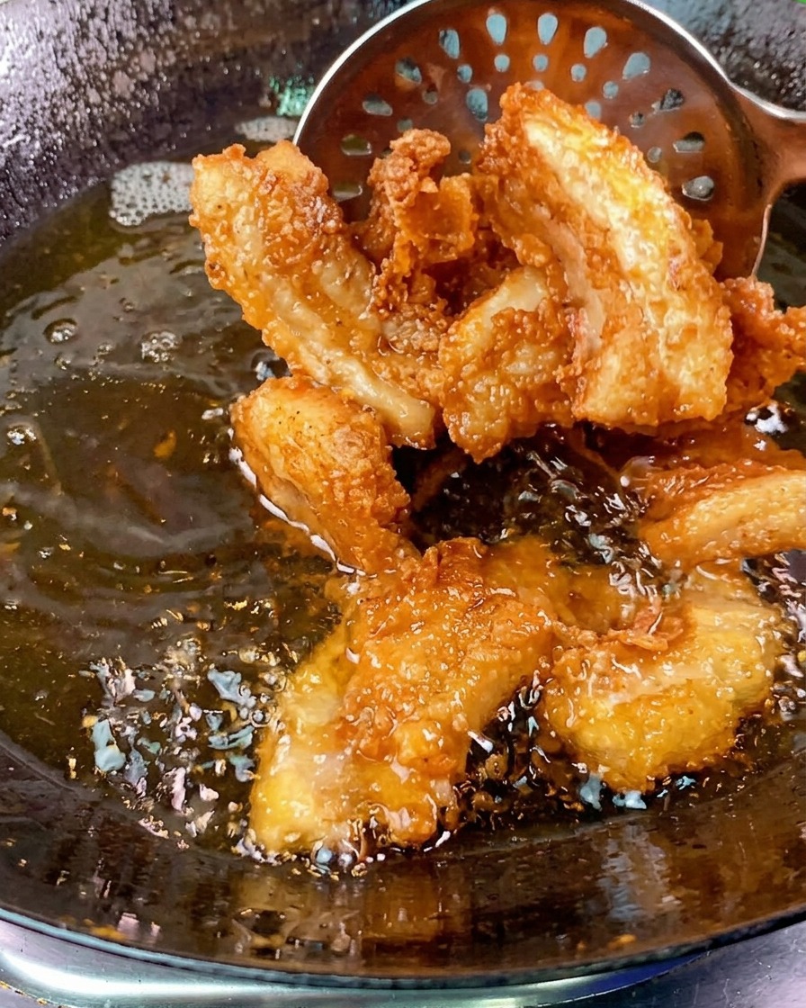 Knusprige goldbraune Schweinebauchscheiben werden mit einer Schaumkelle aus einem sprudelnden Öl-Wok gehoben.