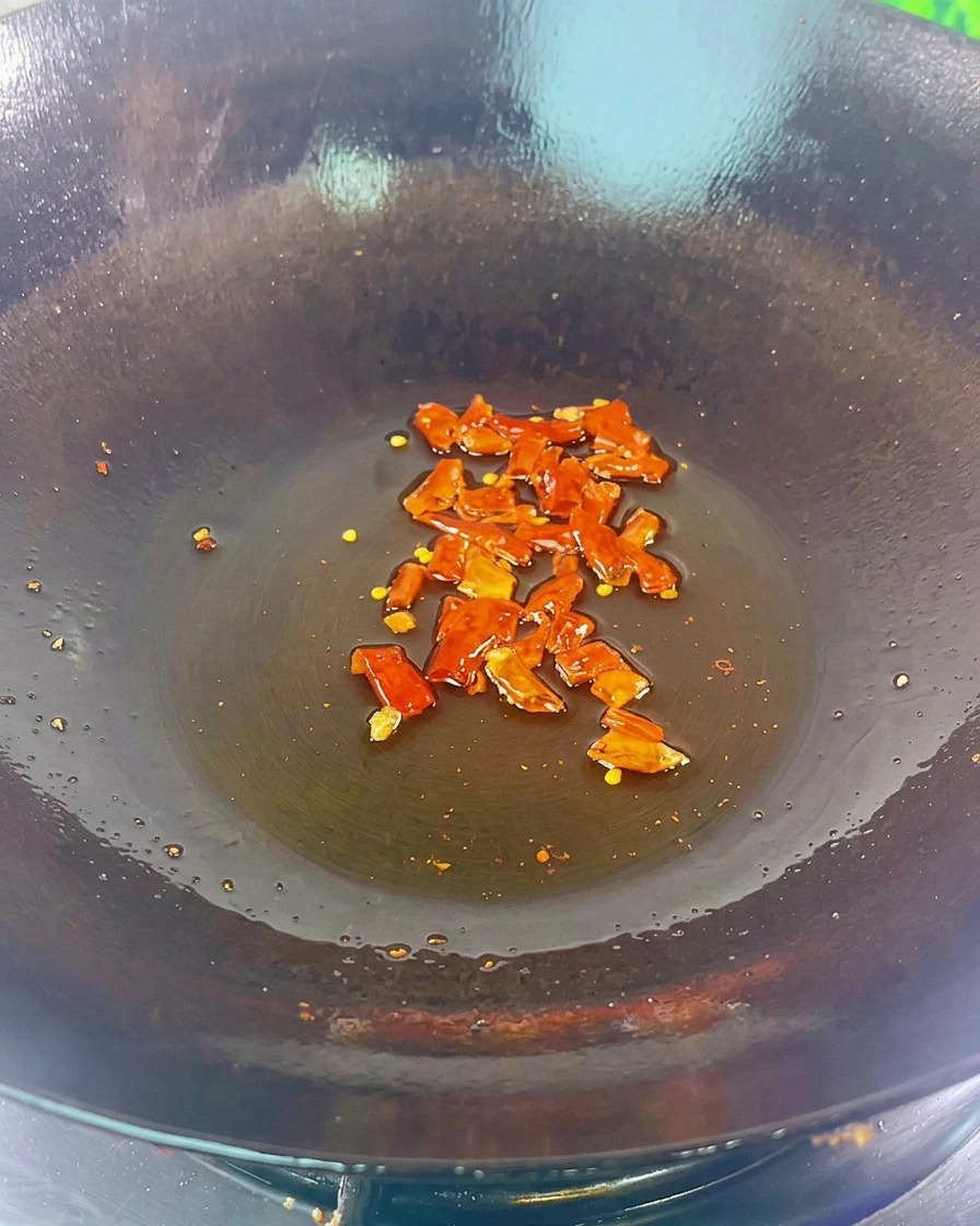 Getrocknete rote Chilis und schwarze Bohnen brutzeln in etwas Öl auf dem Boden eines dunklen Woks.