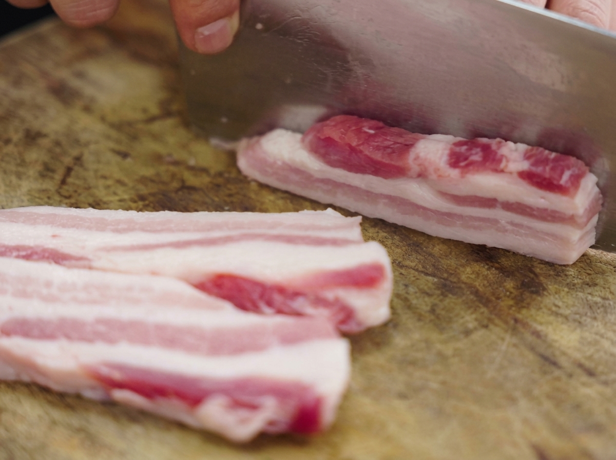 Dickere Streifen von rohem, gleichmäßig geschichtetem Schweinebauch werden mit einem großen Hackmesser auf einem Holzschneidebrett zerteilt.
