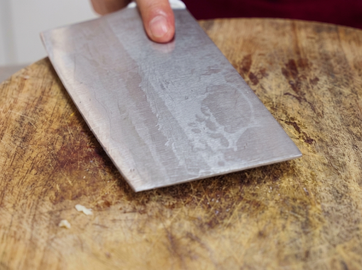 Die flache Seite eines schweren Hackmessers wird auf einem Holzbrett nach unten gepresst, um rohe Knoblauchzehen zu zerdrücken.