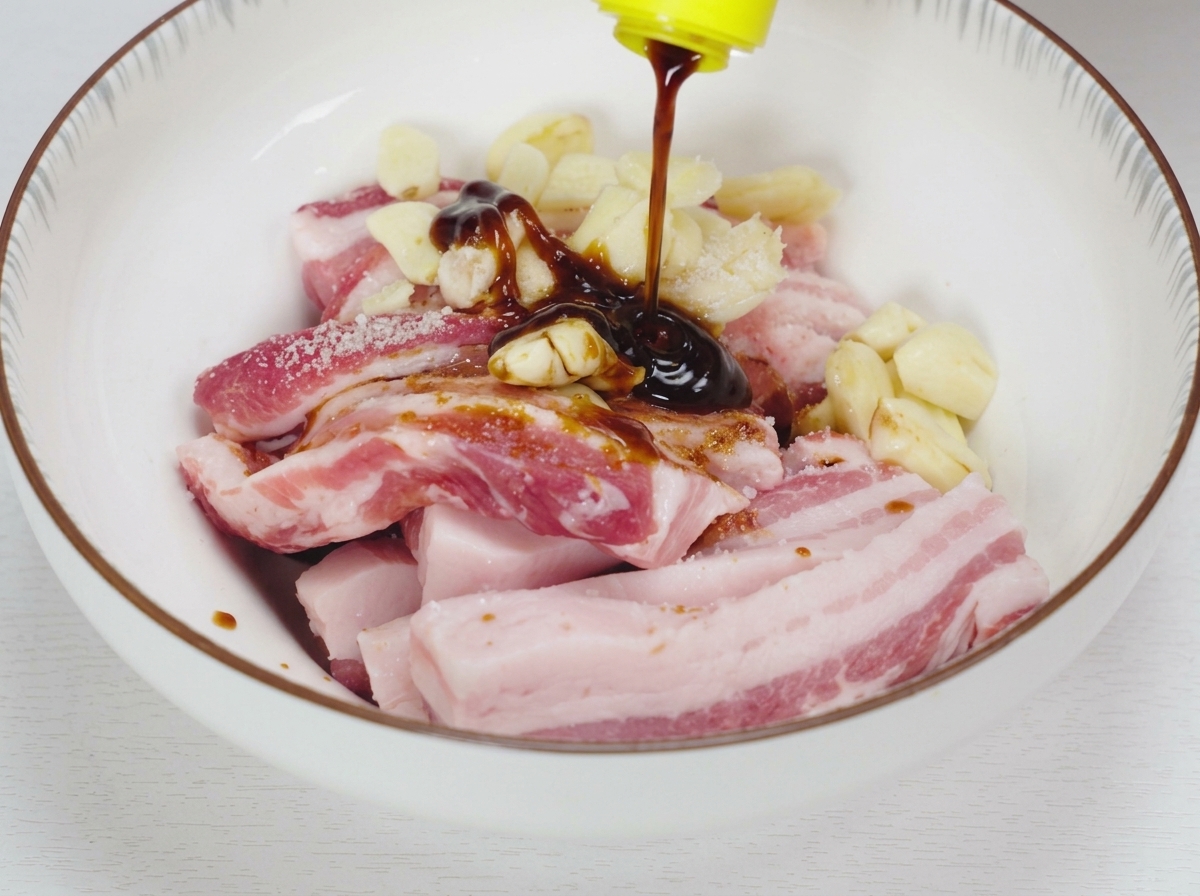 Dunkle flüssige Würzsaucen und Austernsauce werden über rohe Schweinebauchstreifen und zerdrückten Knoblauch in einer weißen Keramikschüssel gegossen.
