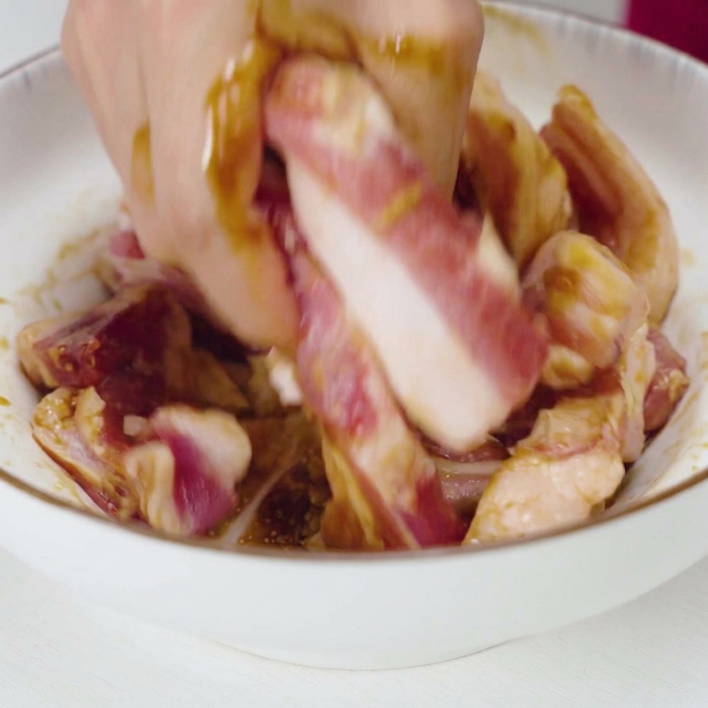 Eine Hand vermengt und knetet rohe Schweinefleischstreifen intensiv mit einer dunklen flüssigen Marinade und Knoblauchstücken in einer weißen Schüssel.