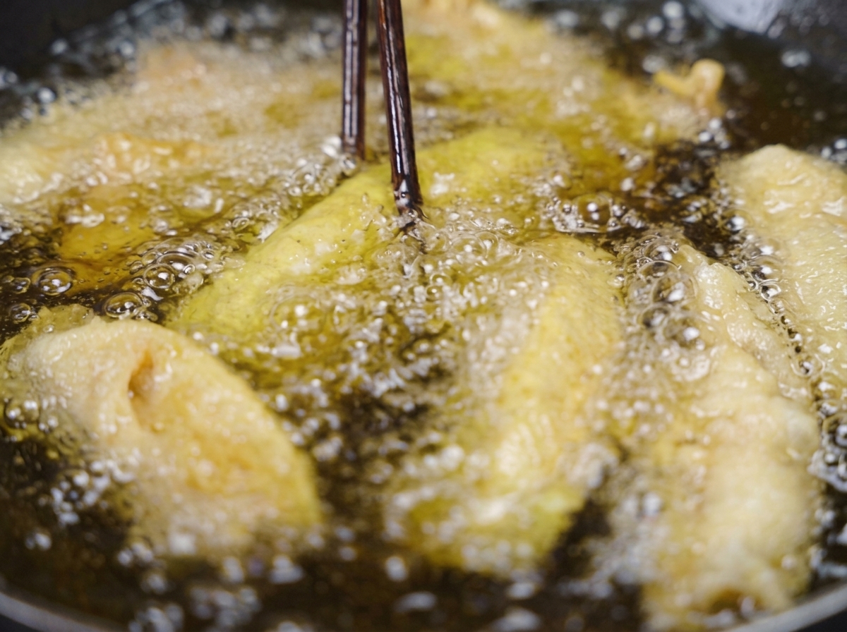 Dunkle Holzstäbchen heben einen knusprigen, goldbraun frittierten Schweinebauchstreifen aus dem sprudelnden Frittieröl.